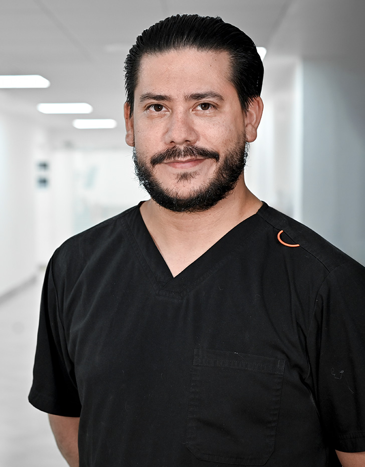 Es la foto de perfil de un cirujano plástico en Guadalajara de Hospital Angeles Del Carmen. Utiliza un uniforme azul marino, tiene barba y bigote negro. Al fondo se ve el hospital.