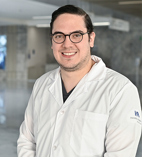 Es la foto de perfil de un angiólogo y cirujano vascular en Querétaro de Hospital Angeles Centro Sur. Usa una bata blanca, una playera negra, está sonriendo y usa lentes.