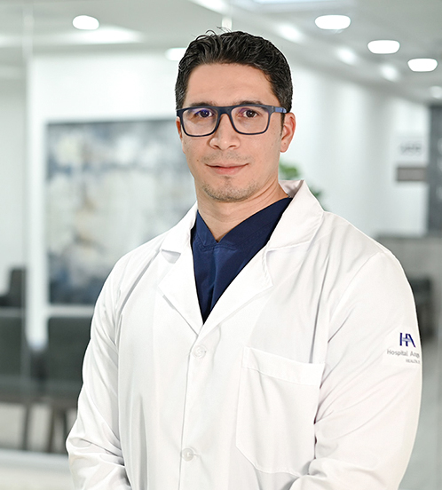 Es la foto de perfil de un cirujano oncólogo en Ciudad de México de Hospital Angeles Acoxpa. Usa una bata blanca con un uniform médico color azul oscuro. Tiene el cabello nehro y usa lentes azules cuadrados.