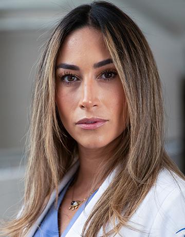 Es la foto de perfil de una angióloga y cirujana vascular y endovascular en Huixquilucan de Hospital Angeles Lomas. Usa una bata blanca, un uniforme azul con collar, cabello largo lacio castaño con luces rubias.