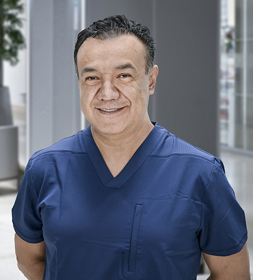 Es la foto de perfil de un cirujano general en Huixquilucan de Hospital Angeles Lomas. Usa un uniforme azul, es de tez morena con cabello oscuro, está de brazos cruzados y está sonriendo.