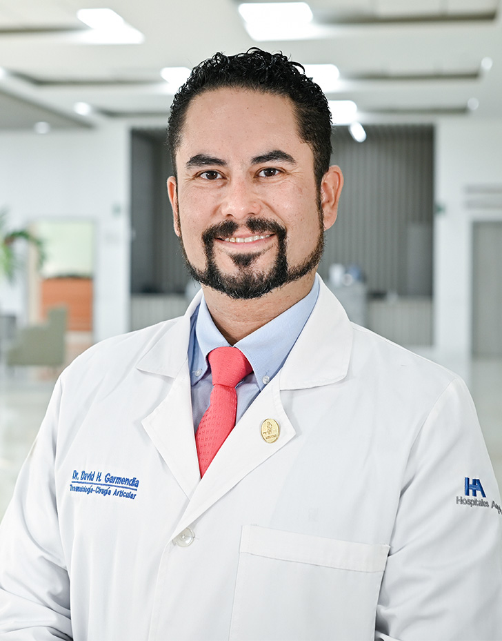 Es la foto de perfil de un ortopedista y traumatólogo en Tampico de Hospital Angeles Tampico. Usa una bata blanca y una corbata roja. Tiene el cabello y barba negra. Tiene los brazos cruzados.