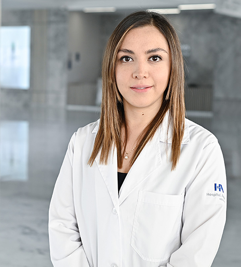 Es la foto de perfil de una algóloga en Querétaro de Hospital Angeles Centro-sur. Usa una bata blanca con una blusa azul oscuro y accesorios plata. Tiene el cabello lacio y castaño. Sus ojos están delineados.