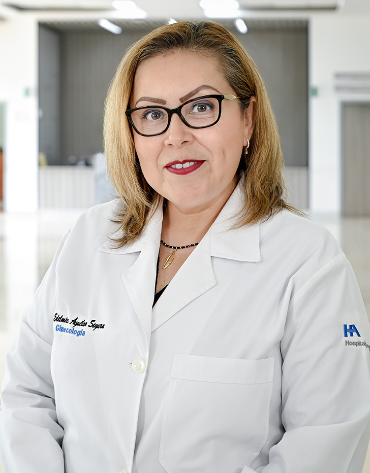 Es la foto de perfil de una ginecóloga y obstetra en Tampico de Hospital Angeles Tampico. Usa una bata blanca y una blusa gris. Tiene el cabello corto y chino