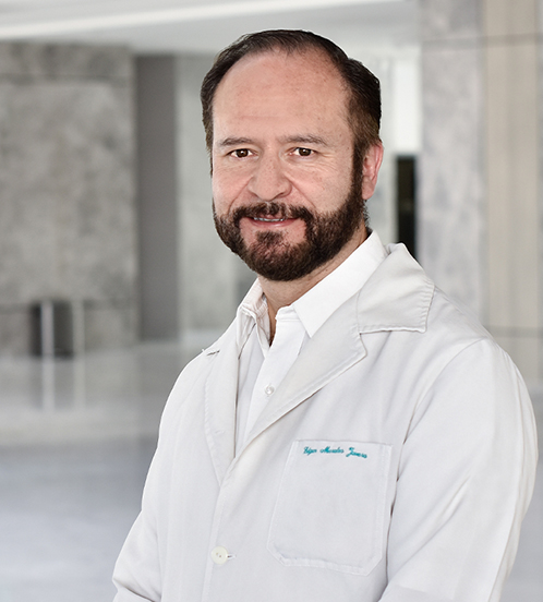 Es la foto de perfil de un urólogo pediátrico en Querétaro de Hospital Angeles Centro Sur. Usa una camisa blanca con una bata blanca, tiene barba y está sonriendo.