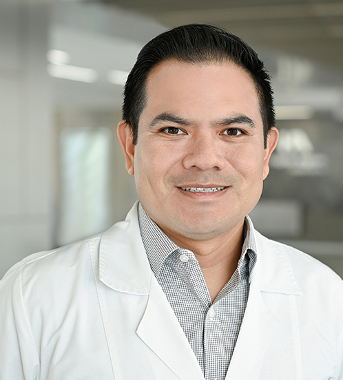 Es la foto de perfil de un nefrólogo pediátrico en Querétaro de Hospital Angeles Centro Sur. Usa una bata blanca con una camisa blanca con puntos negros, está sonriendo.