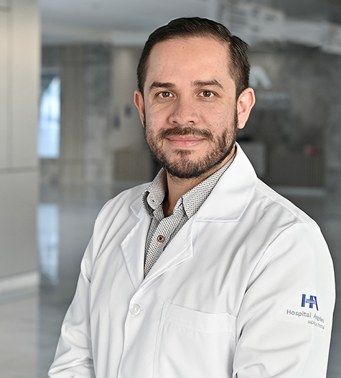 Es la foto de perfil de un oncólogo médico en Querétaro de Hospital Angeles Centro-sur. Tiene cabello, barba y ojos de color café. Usa una bata médica blanca y una camisa blanca con detalles grises.