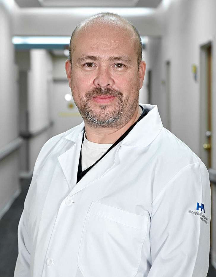 Es la foto de perfil de un cirujano cardiotorácico en Torreón de Hospital Angeles Torreón. Usa una bata blanca y una camisa azul claro. Es calvo y tiene barba café.