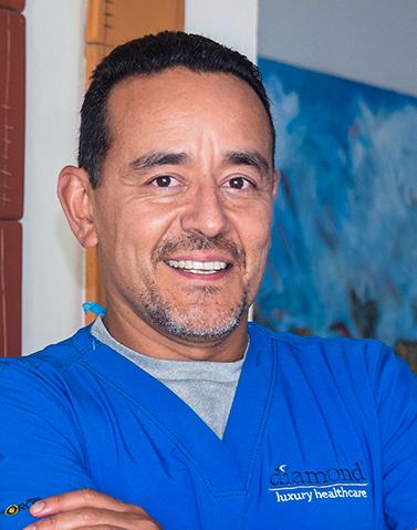 Es la foto de perfil de un cirujano oncológico en Xalapa de Hospital Angeles Xalapa. Usa un uniforme médico color azul. Tien el cabello y barba negra. Tiene los brazos cruzados.