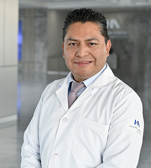 Es la foto de perfil de un cirujano general en Querétaro de Hospital Angeles Centro-sur. Usa una bata médica color blanco con una camisa azul claro y una corbata gris.  Está sonriendo ligeramente y tiene el cabello negro con canas.