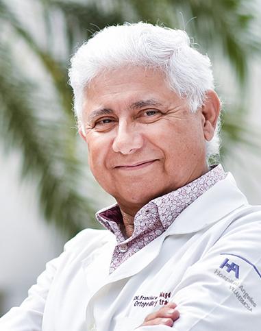 Es la foto de perfil de un ortopedista y traumatólogo en Villahermosa de Hospital Angeles Villahermosa. Usa una bata blanca con una camisa rosada. Tiene le cabello blanco y los brazos cruzados.