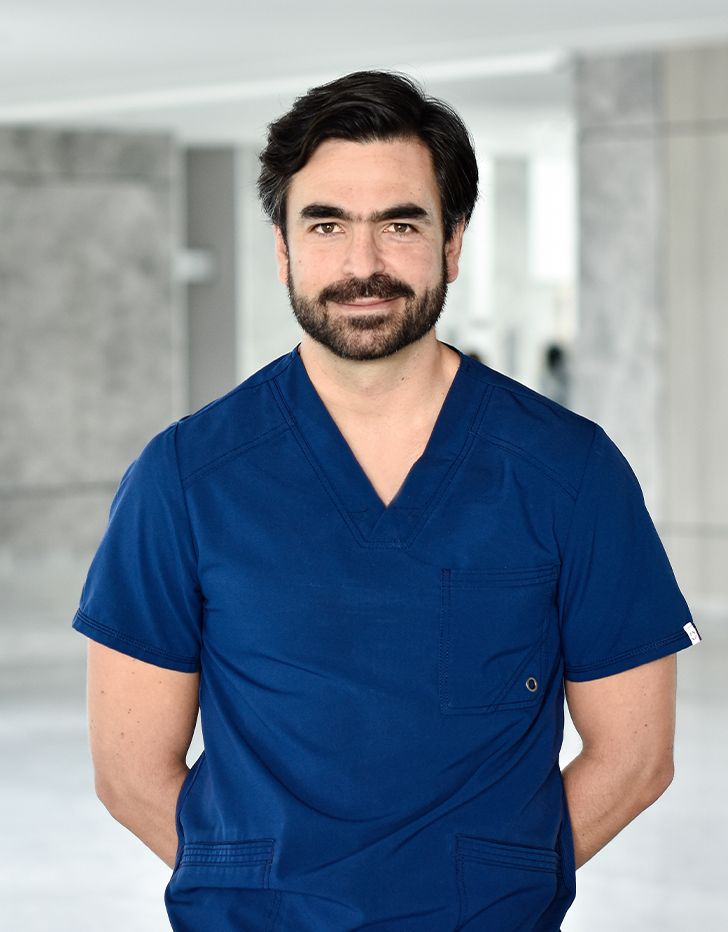 Es la foto de perfil de un ortopedista, traumatólogo y especialista en artroscopía en Querétaro de Hospital Angeles Querétaro. Usa un uniforme médico azul. Tiene el cabello y barba negra. Tiene los brazos cruzados por detrás.