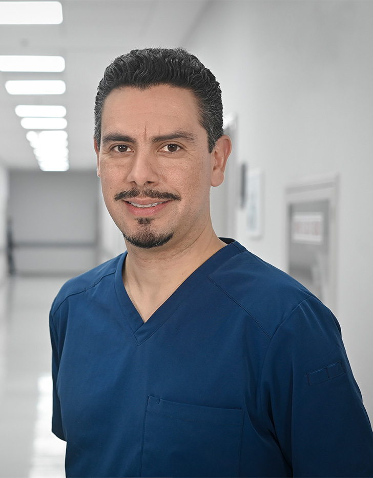 Es la foto de perfil de un angiólogo y cirujano vascular en Ciudad de México de Hospital Angeles Universidad. Usa un uniforme médico azul. Tiene barba y cabello negro.