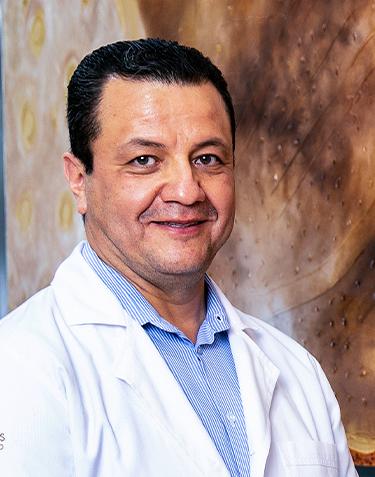 Es la foto de perfil de un ginecólogo y obstetra en León de Hospital Angeles León. Utiliza una bata con camisa azul, de fondo hay una imagene n colores café oscuro, beige y amarillo.