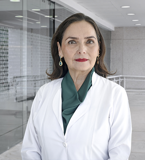 Es la foto de perfil de una ginecóloga y obstetra en Villahermosa de Hospital Angeles Villahermosa. Usa una bata blanca y una blusa con estampados color morada. Tiene el cabello corto de color café. Usa labial rojo y está sonriendo.