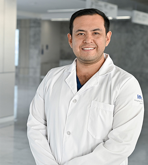 Es la foto de perfil de un pediatra en Querétaro de Hospital Angeles Centro-sur. Se encuentra de pie con las manos cruzadas por enfrente. Usa una bata blanca y un uniforme médico azul. Está sonriendo.