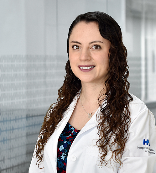 Es la foto de perfil de una ginecóloga y obstetra en Huixquilucan de Hospital Angeles Lomas. Usa una bata blanca con una blusa negra con azul claro. Su cabello es largo y rizado, está sonriendo.