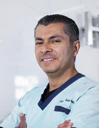 Es la foto de perfil de un nefrólogo en Ciudad de México de Hospital Angeles. El doctor está sonriendo y viste un uniforme de color menta y cuello azul marino.