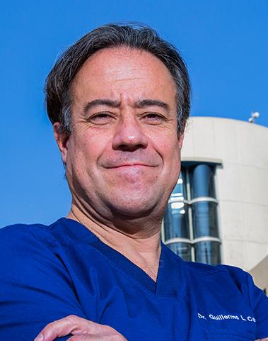 Es la foto de perfil de un ginecólogo y obstetra en Huixquilucan de Hospital Angeles Lomas. Usa un uniforme azul, tiene el cabello ligeramente largo, tiene los brazos cruzados y está sonriendo.