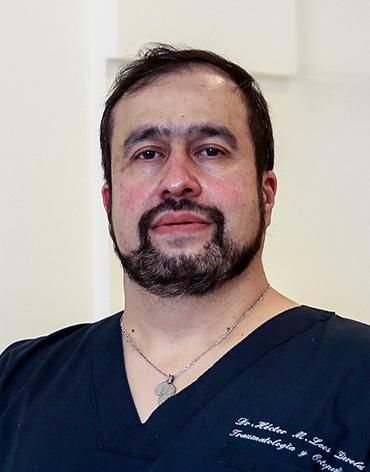 Es la foto de perfil de un ortopedista y traumatólogo en León de Hospital Angeles León. Usa un uniforme azul marino en cuello V tiene un collar, barba y bigote.