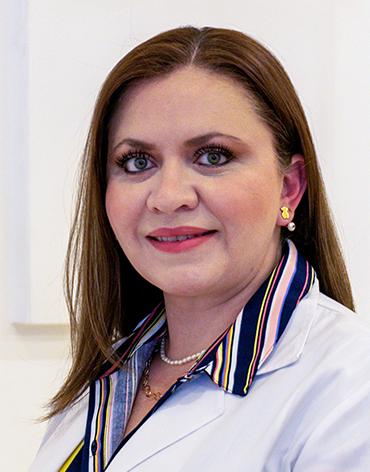 Es la foto de perfil de una ginecóloga y obstetra en León de Hospital Angeles León. Utiliza una bata blanca con una camisa azul marino y líneas. Tiene el cabello castaño claro.