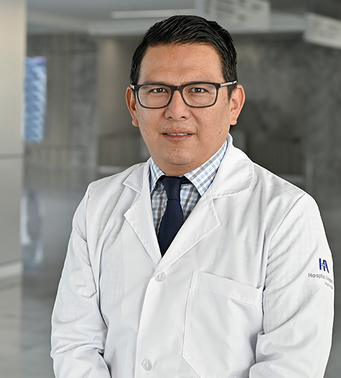 Es la foto de perfil de un cirujano oncológico en Querétaro de Hospital Angeles Centro Sur. Usa un uniforme azul, tiene una cadena dorada y lentes con marco negro.