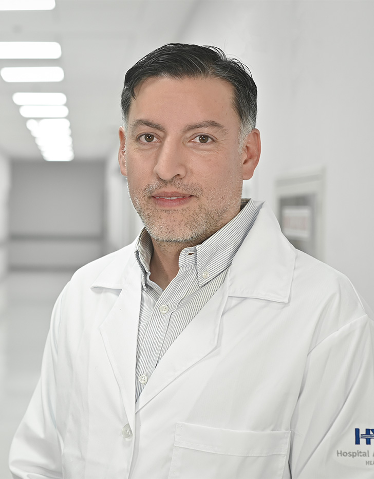 Es la foto de perfil de un urólogo en Ciudad de México de Hospital Angeles Universidad. Usa una bata blanca y una camisa color gris. Tiene el cabello negro.