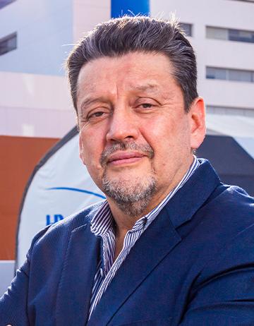 Es la foto de perfil de un cirujano general en Tijuana de Hospital Angeles Tijuana. Usa un saco azul. Tiene el cabezo negro grisaseo y barba. Tiene los brazos cruzados.