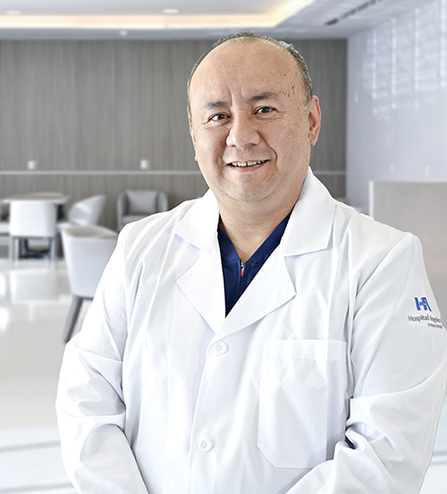 Es la foto de perfil de un cirujano plástico en San Pedro Garza de Hospital Angeles Valle Oriente. Usa una bata blanca con una camisa azul claro y una corbata vino. Está sonriendo y está sentado.