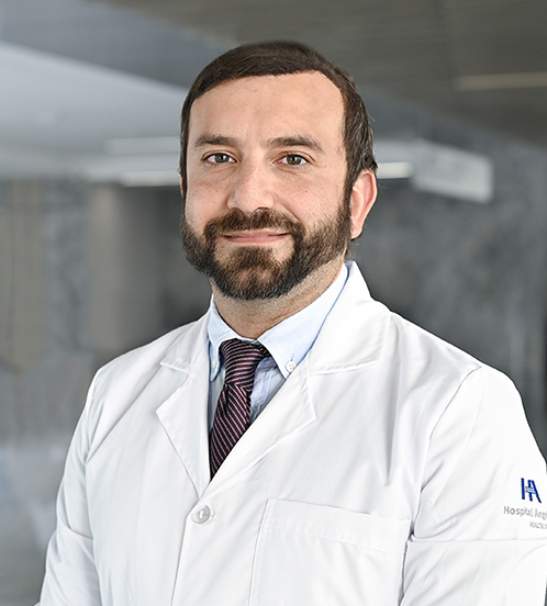 Es la foto de perfil de un oncólogo médico en Querétaro de Hospital Angeles Centro Sur. Usa una bata y una camisa de rayas color azul, tiene barba y de fondo es la recepción del hospital.