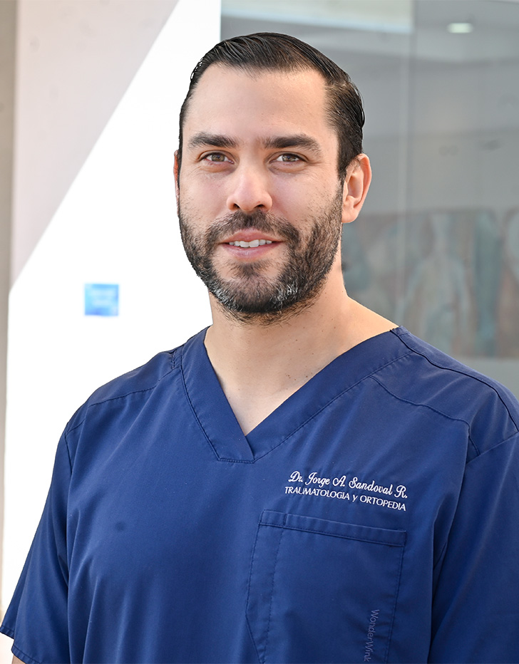 Es la foto de perfil de un ortopedista y traumatólogo en Tijuana de Hospital Angeles Tijuana. Usa un uniforme médico color azul. Tiene el cabello y barba café oscuro. Está sonriendo.