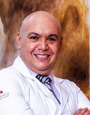 Es la foto de perfil de un cirujano general en León de Hospital Angeles León. Utiliza una bata blanca con una camisa clara y una corbata. Tiene los brazos cruzados y está sonriendo, es calvo.