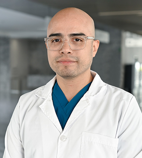 Es la foto de perfil de un gastroenterólogo en Querétaro de Hospital Angeles Centro-sur. Usa una bata blanca con un uniforme médico azul. Es calvo y usa lentes con armazón transparente.