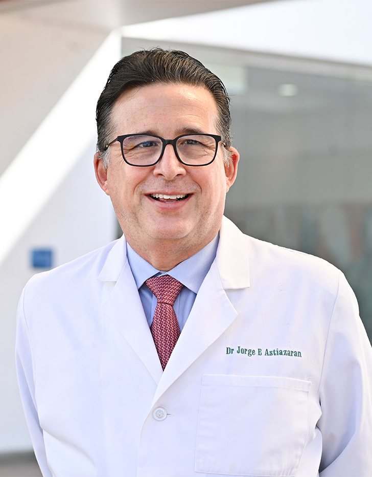 Es la foto de perfil de un internista en Tijuana de Hospital Angeles Tijuana. Usa una bata blanca con una camisa azul y corbata roja. Tiene el cabello negro y usa lentes. Está sonriendo.