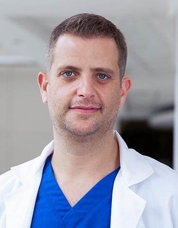 Es la foto de perfil de un ortopedista y traumatólogo en Huixquilucan de Hospital Angeles Lomas. El doctor usa una bata blanca, tiene un uniforme médico azul y los ojos azules.