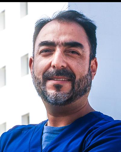 Es la foto de perfil de un neurólogo en Torreón de Hospital Angeles Torreón. Usa un uniforme médico azul. Tiene la barba y cabello negro con gris. Está sonriendo y tiene los brazos cruzados.