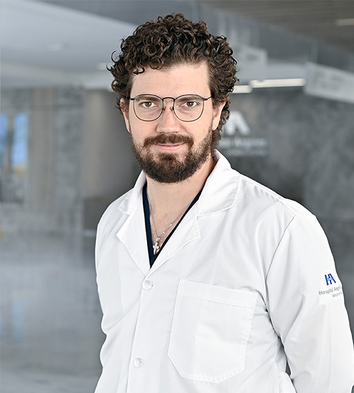 Es la foto de perfil de un cardiólogo en Querétaro de Hospital Angeles Centro-sur. Usa una bata blanca con un uniforme médico color azul oscuro. También usa una cadena de cruz color plata. Tiene el cabello castaño y chino.