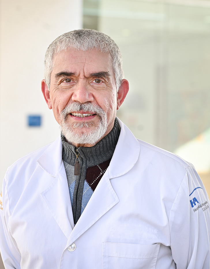 Es la foto de perfil de un pediatra e infectólogo pediátrico en Tijuana de Hospital Angeles Tijuana. Usa una bata blanca y un sueter color negro con gris. Tiene el cabello y baraba blanca. Está sonriendo.