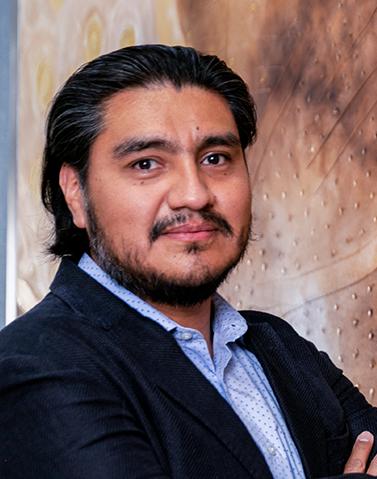 Es la foto de perfil de un cirujano oncológico en León de Hospital Angeles León. Tiene un sco negro, una camisa azul cielo, el cabello largo, tiene bigote y barba.