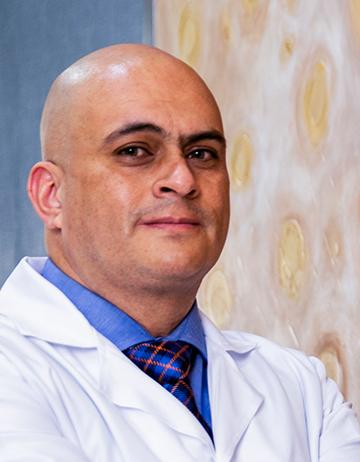 Es la foto de perfil de un reumatólogo en León de Hospital Angeles León. Usa una bata blanca con una camisa azul y una corbata, es calvo y tiene cejas pobladas.