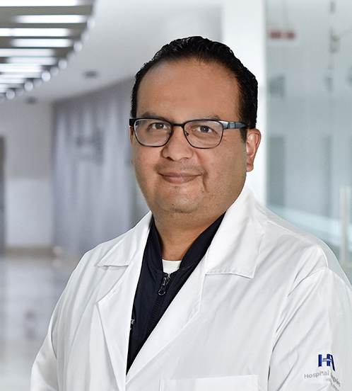 Es la foto de perfil de un angiólogo y cirujano vascular en Huixquilucan de Hospital Angeles Lomas. Usa una bata blanca y una playera negra, tiene lentes cuadrados y está sonriendo.