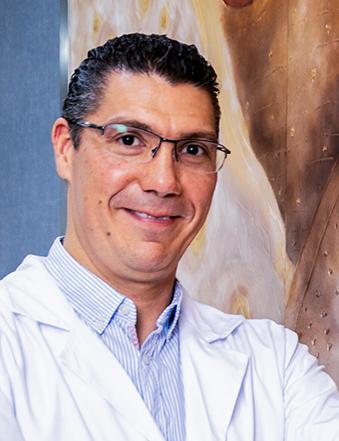 Es la foto de perfil de un gastroenterólogo especialista en gastrocirugía y cirugía general en León de Hospital Angeles León. Tiene una bata blanca con una camisa azul cielo, tiene lentes y está sonriendo. 