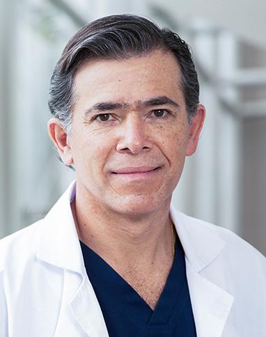 Es la foto de perfil de un cirujano maxilofacial en Huixquilucan de Hospital Angeles Lomas. Usa una bata blanca con un uniforme negro. está sonriendo.