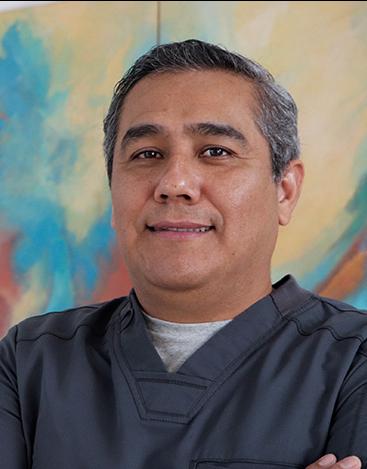 Es la foto de perfil de un cirujano general en Tampico de Hospital Angeles. Usa un uniforme médico gris. Tiene los brazos cruzados y una sonrisa ligera.