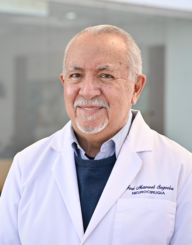 Es la foto de perfil de un cirujano neurológico en Tijuana de Hospital Angeles. Usa una bata blanca con un sueter azul y una camisa azul claro. Tiene el cabello y bigote gris. Está sonriendo.