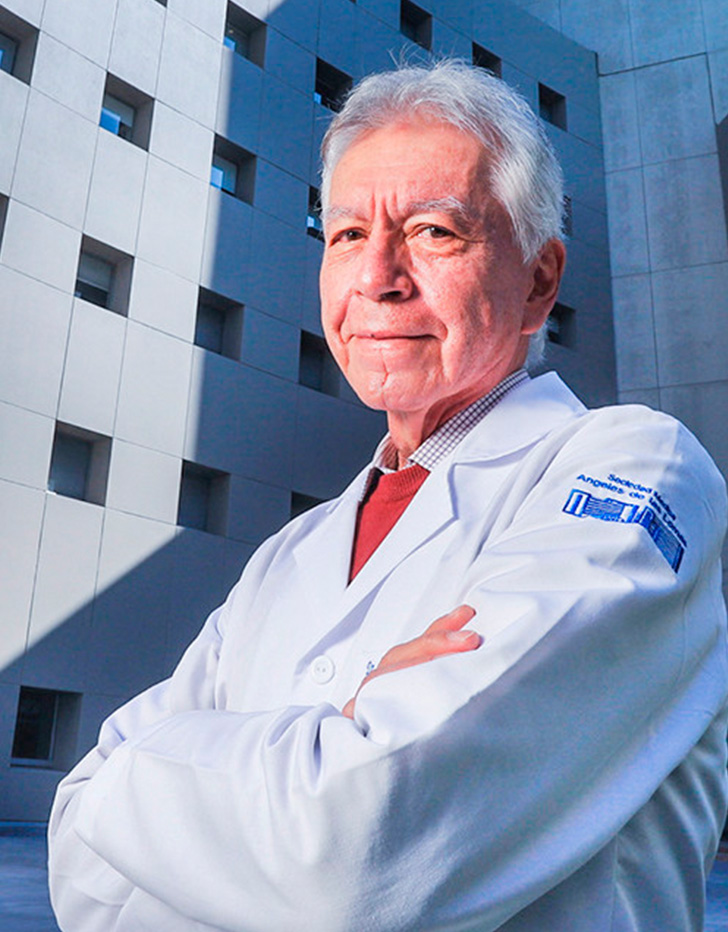 Es la foto de perfil de un ortodoncista en Huixquilucan de Hospital Angeles Lomas. Usa bata blanca, tiene el cabello canoso y los brazos cruzados. 