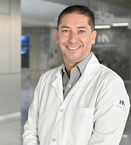 Es la foto de perfil de un ginecólogo y obstetra en Querétaro de Hospital Angeles Centro-sur. Usa una bata blanca con una camisa gris. Tiene el cabello negro. Tiene los brazos cruzados en la parte de enfrente.