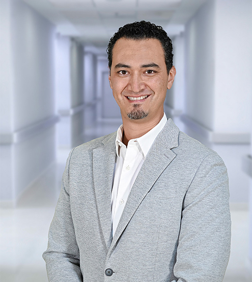 Es la foto de perfil de un angiólogo y cirujano vascular en Ciudad Juárez de Hospital Angeles Ciudad Juárez. Trae puesta una bata blanca y una playera en cuello V azul marino, el doctor sonríe suavemente.