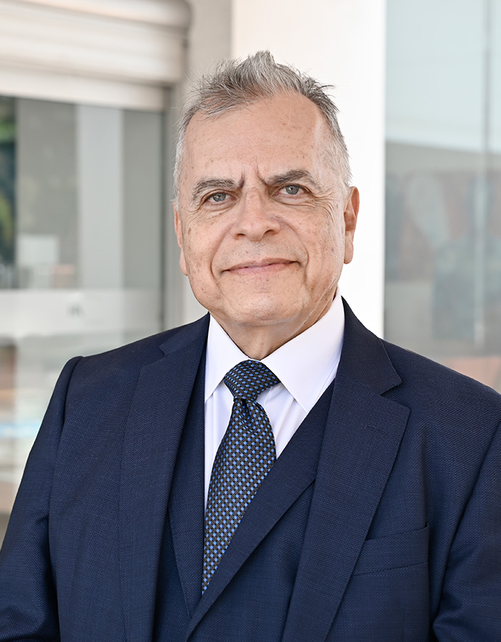 Es la foto de perfil de un cirujano general en Tijuana de Hospital Angeles. Usa un saco negro con camisa blanca y corbata azul. Tiene el cabello gris. Está sonriendo.