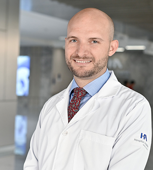 Es la foto de perfil de un ortopedista y traumatólogo en Querétaro de Hospital Angeles Centro-sur. Es calvo. Usa una bata blanca, camisa azul y una corbata roja con detalles azules y está sonriendo.